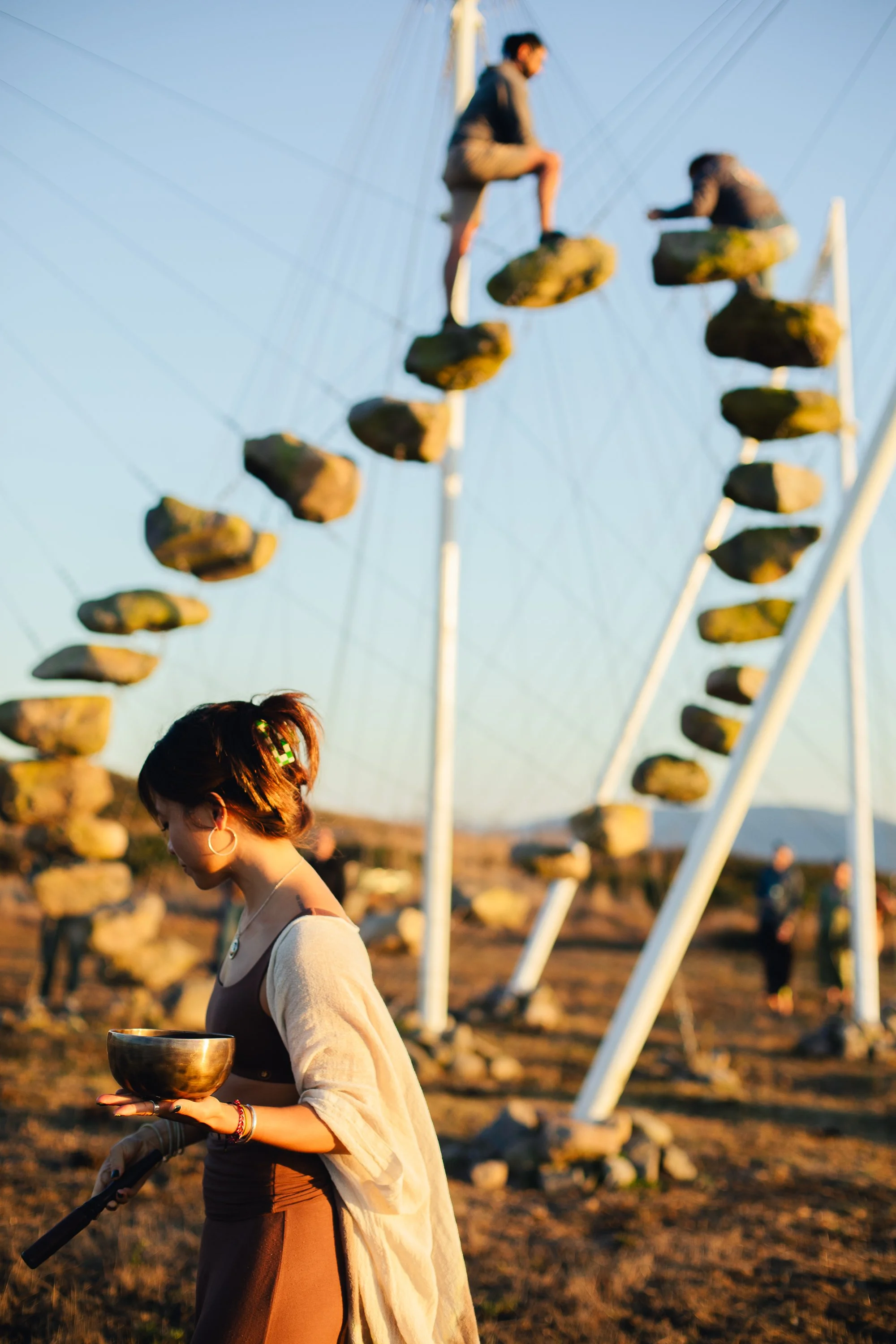 Erica Nguyễn holding a singing bowl in front of a large suspended-stone sculpture installation at golden hour