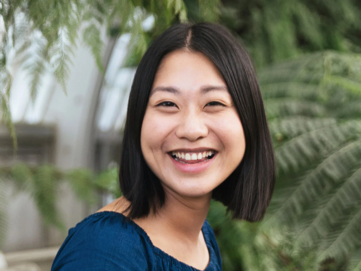 Portrait of Julisa Lau, a smiling woman with a straight black bob in a blue top with greenery behind her
