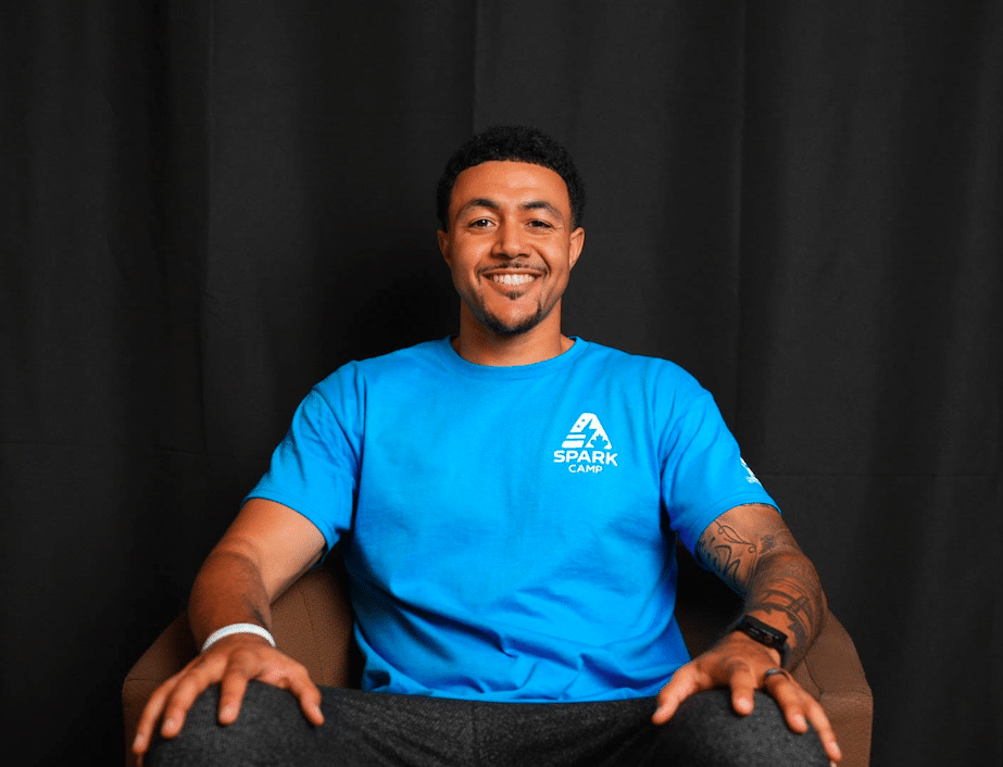 Dom Johnson in a blue Spark Camp t-shirt seated and smiling