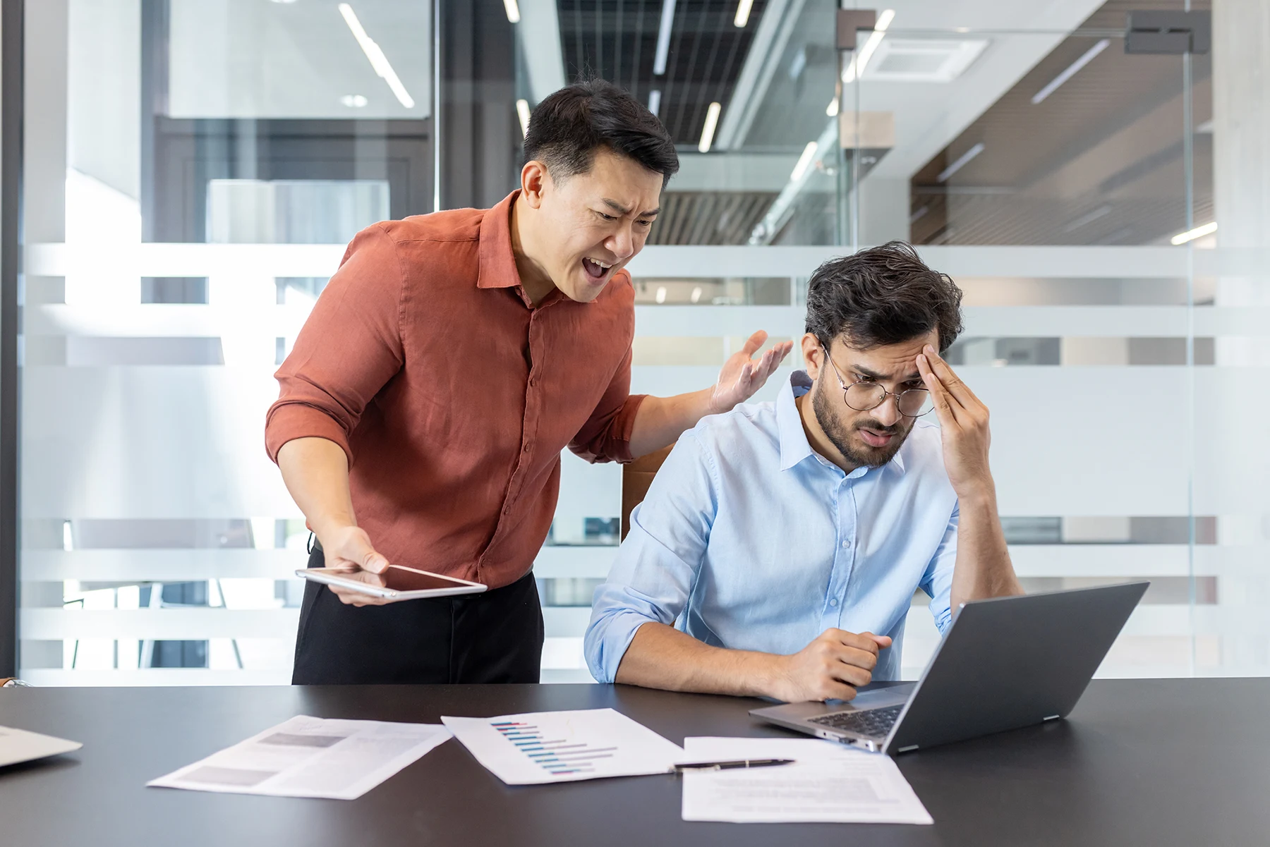 A manager shouting at a stressed colleague working at a laptop