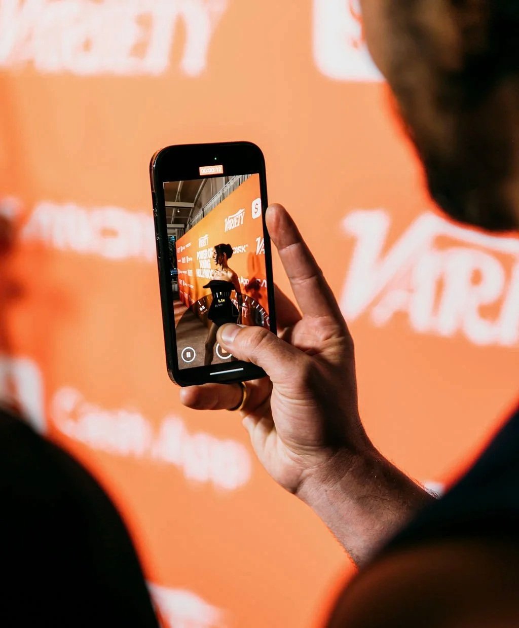 Person holding a phone photographing a celebrity at the Variety orange step-and-repeat backdrop