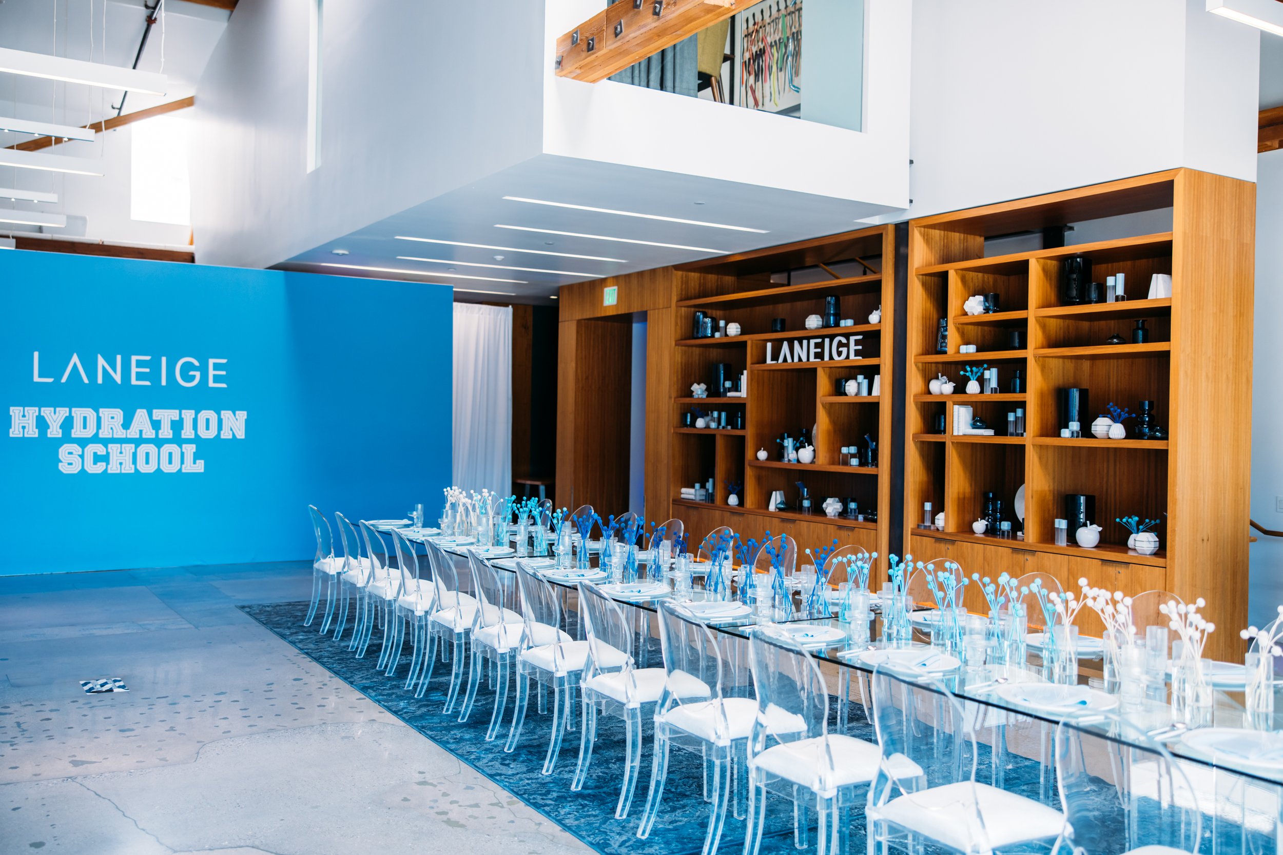 Laneige Hydration School event interior with long glass table, clear ghost chairs, blue branded backdrop wall and wood product shelves
