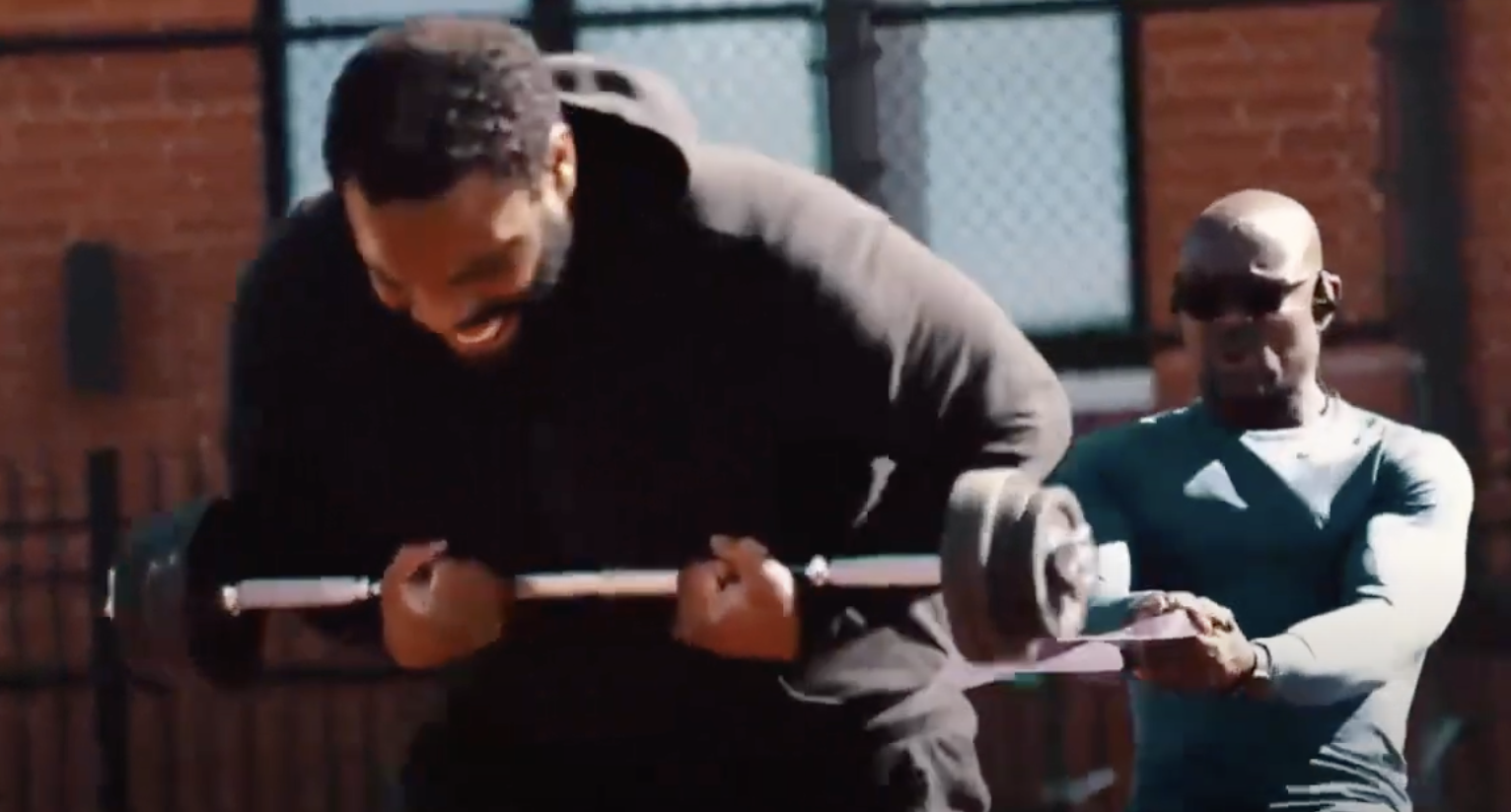 Sunny Akhigbe spotting a client through a barbell curl against an exposed brick gym wall