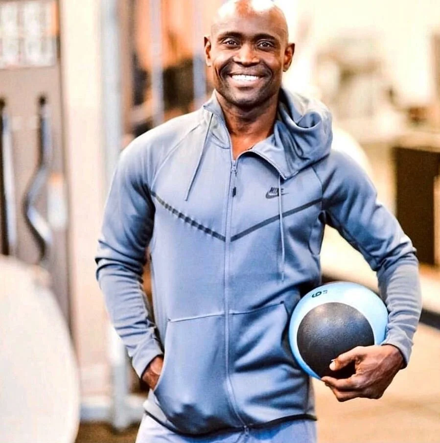 Sunny Akhigbe smiling in a light blue hoodie, holding a medicine ball in a gym