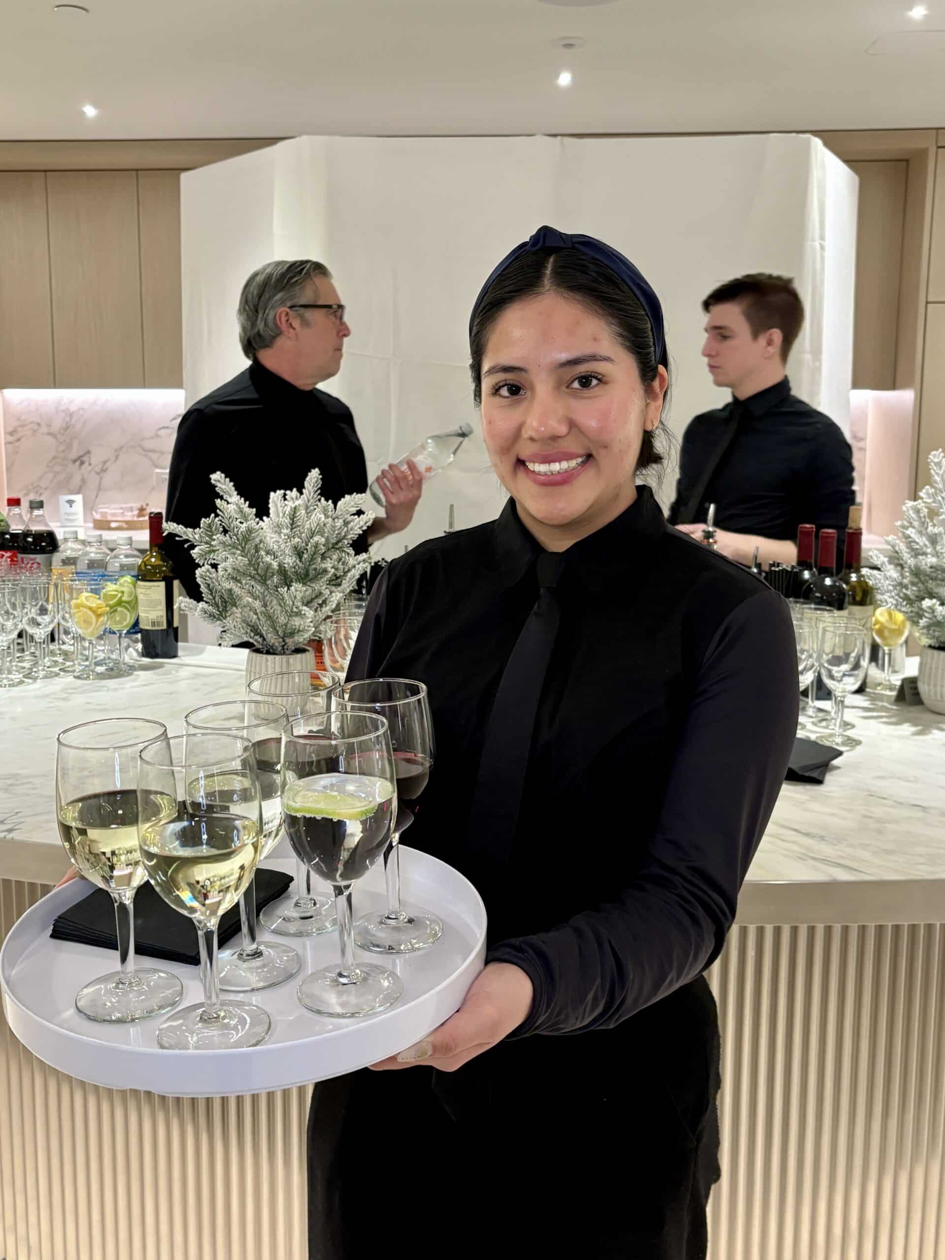 Smiling server in black holding a tray of white wine glasses in front of a modern event bar with two bartenders working behind her