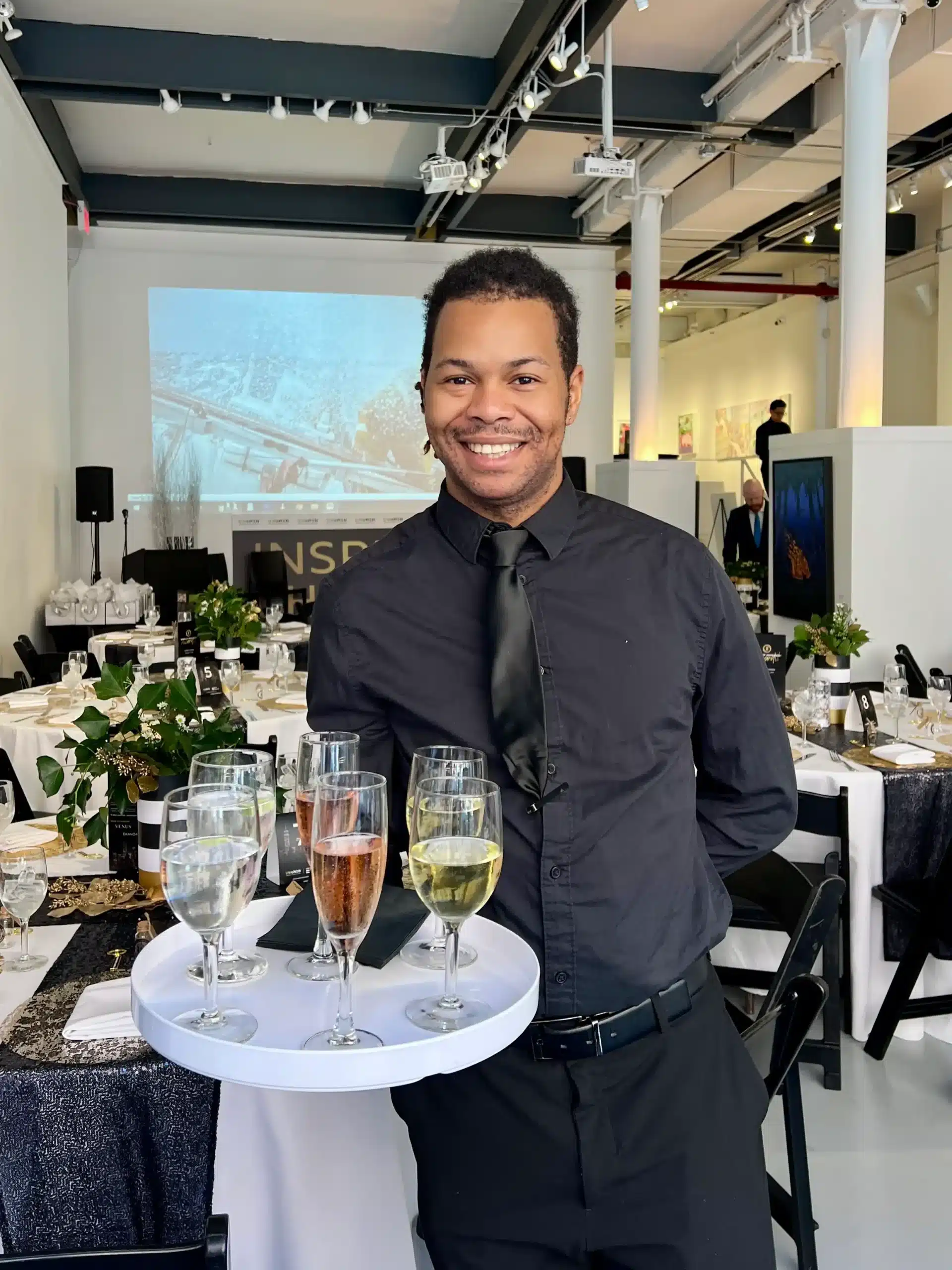 Smiling Sweet Basil server in black shirt and tie holding a tray of champagne and wine at a seated gala