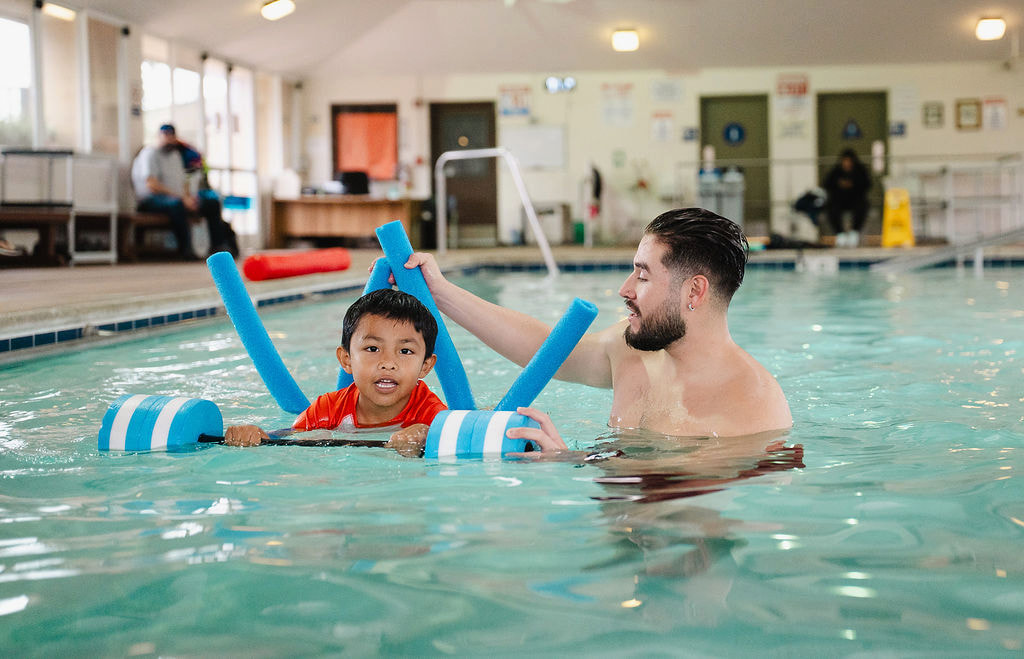A bearded instructor assisting a young boy with foam noodles in an indoor pool