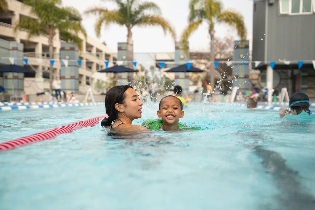 A female instructor holding a laughing toddler in an outdoor pool with palm trees behind