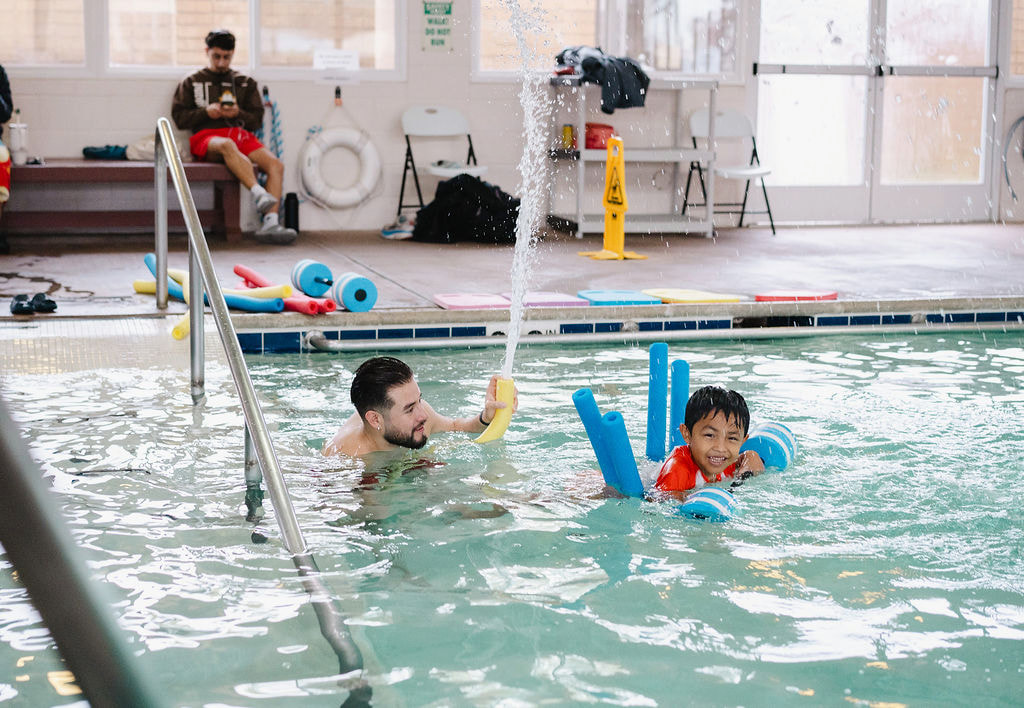 A male swim instructor gently supporting a young boy with foam noodles during an indoor pool lesson