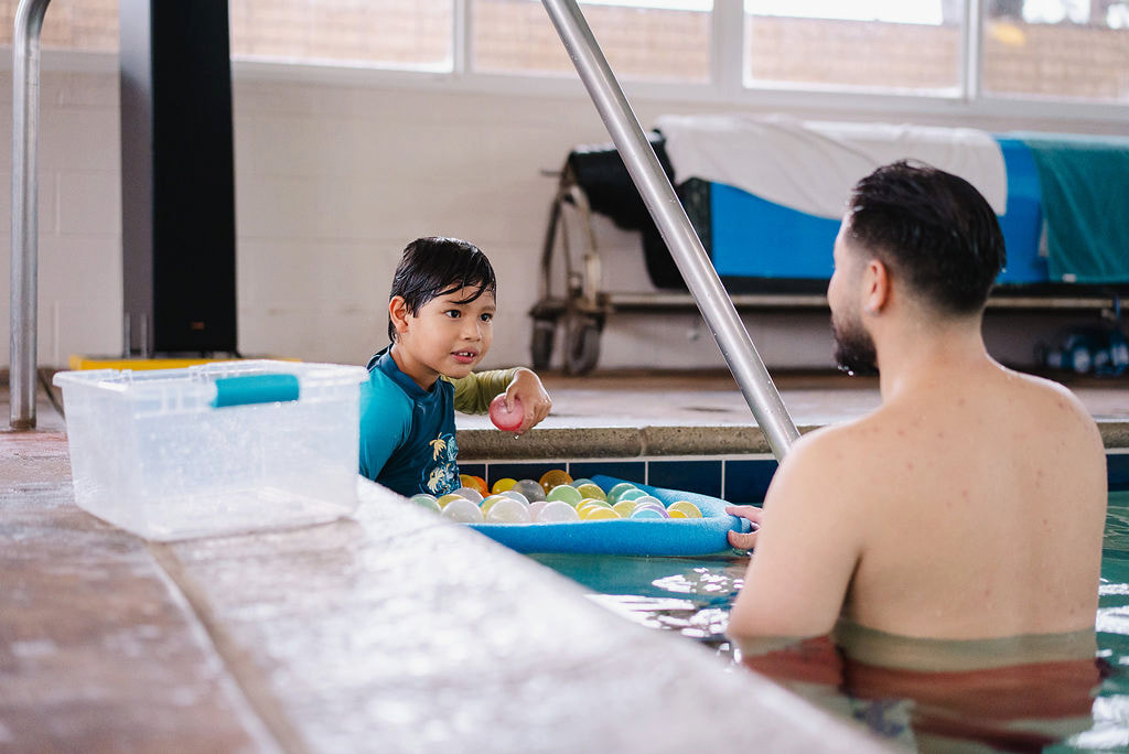 A boy sitting on the pool edge interacting with a bearded male instructor in the water