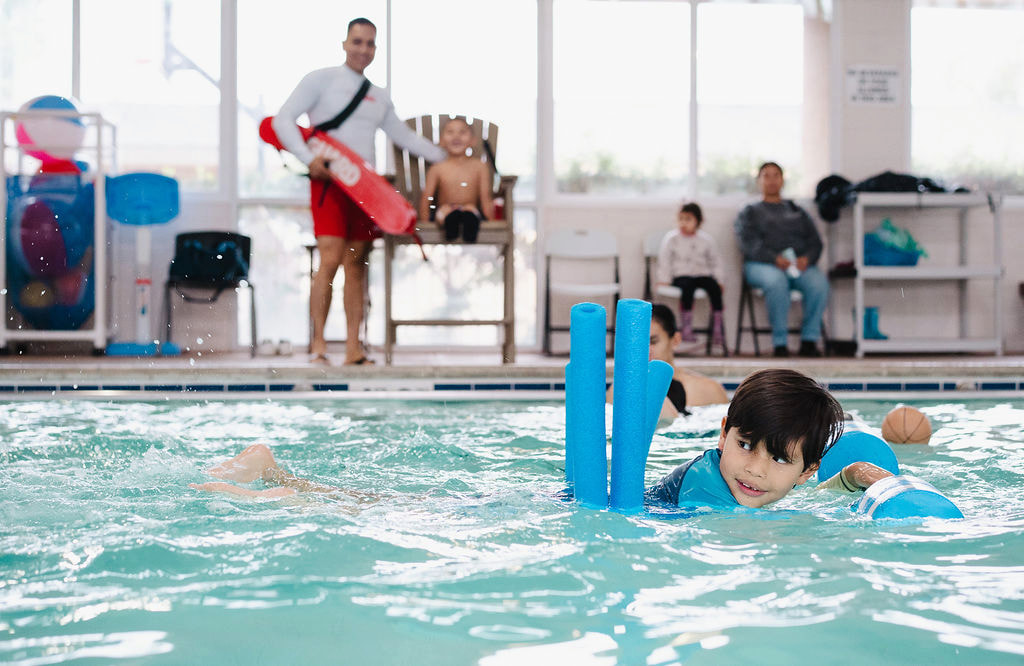 A young boy with foam noodles swimming toward the camera in an indoor pool