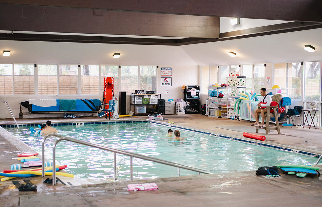Wide shot of the indoor pool facility with a lifeguard, swimmers and equipment shelves