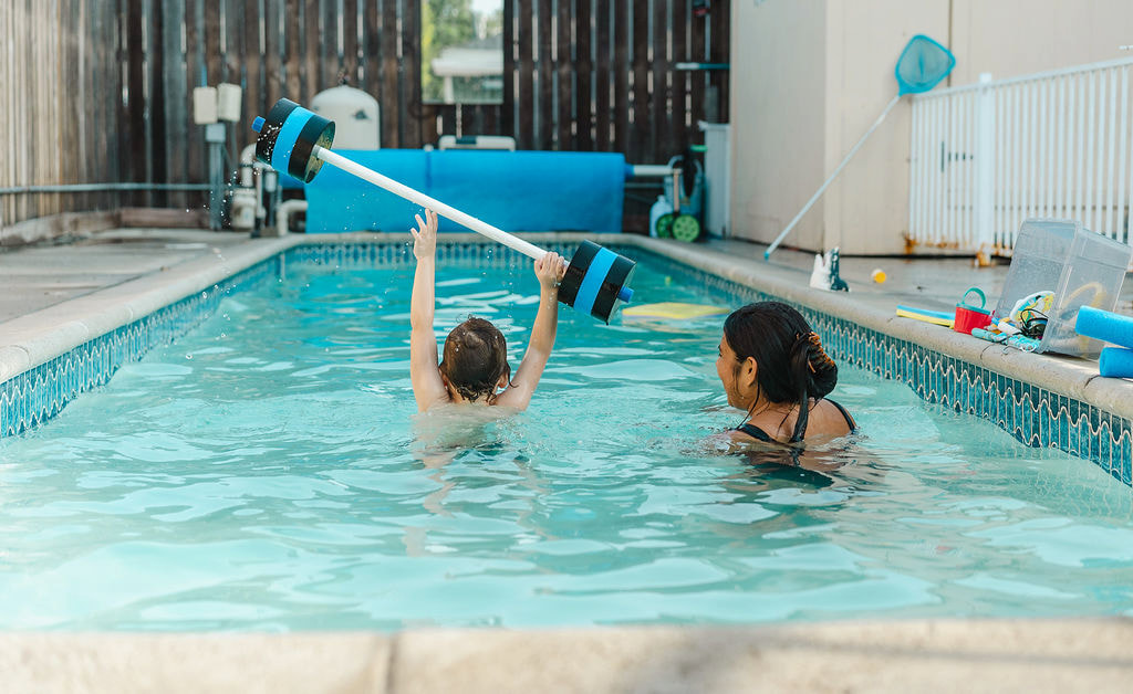 A child lifting a foam barbell overhead while a female instructor watches in a backyard pool