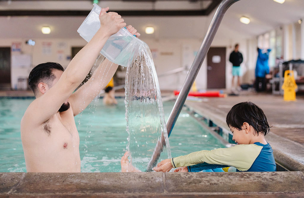 A male instructor pouring water over the pool edge toward a young child indoors
