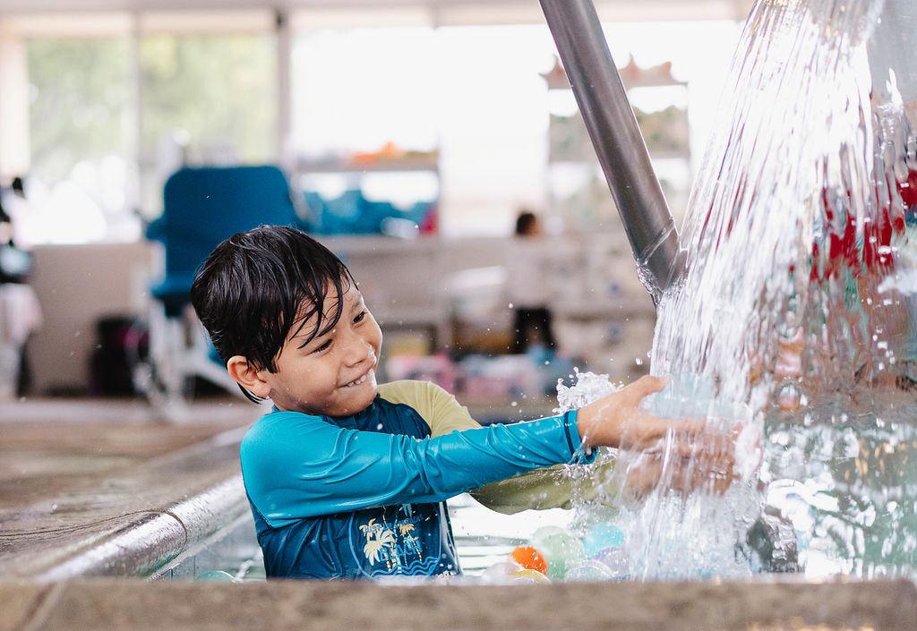 A smiling young boy in a blue rash guard playing with water spray at the pool edge