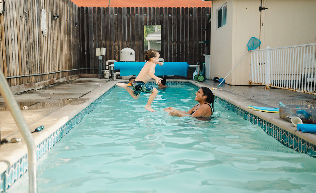 A young child jumping into a backyard pool toward a smiling female instructor