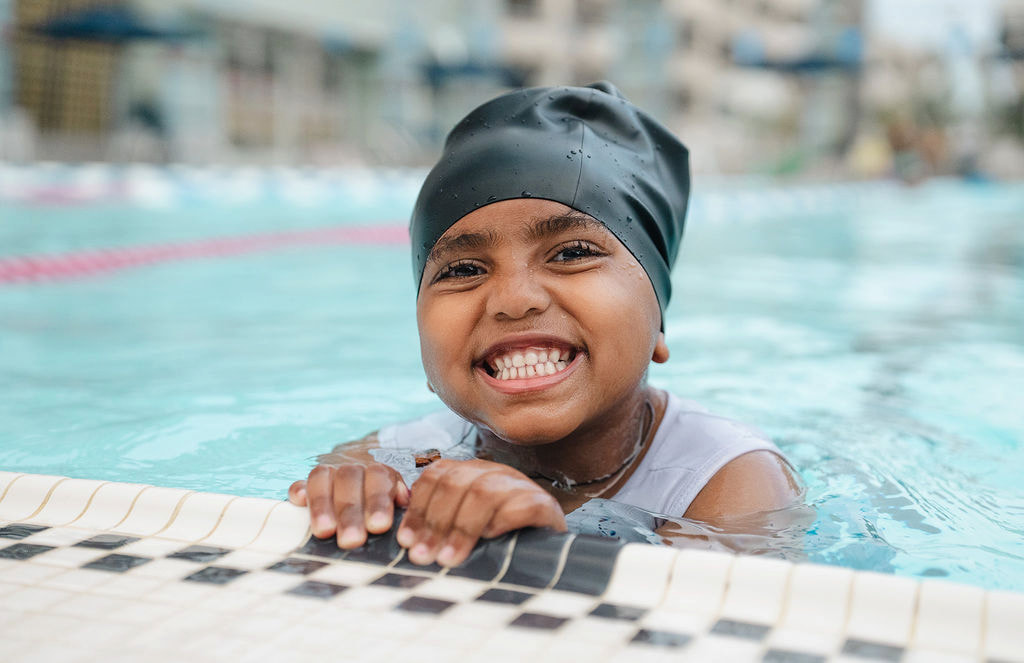 A smiling child in a dark swim cap holding the edge of an outdoor pool