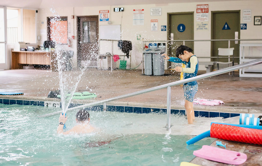 A child at the pool handrail while an instructor splashes water below in an indoor pool