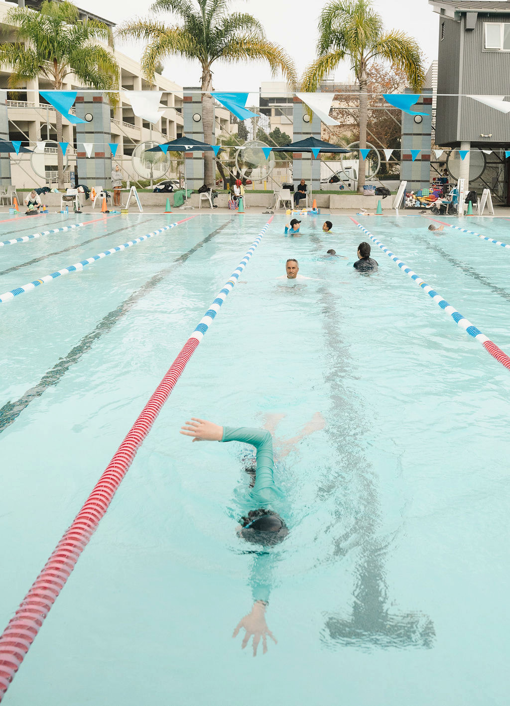 Overhead view of an outdoor lap pool with swimmers, instructors, lane lines and palm trees
