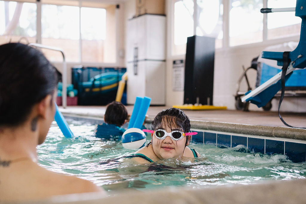 A young girl with pink-accented goggles smiling at the pool edge during an indoor lesson