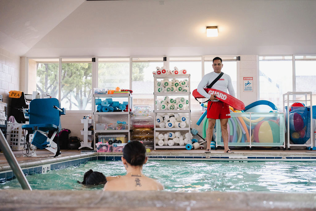 A lifeguard standing poolside holding a rescue buoy at the indoor facility