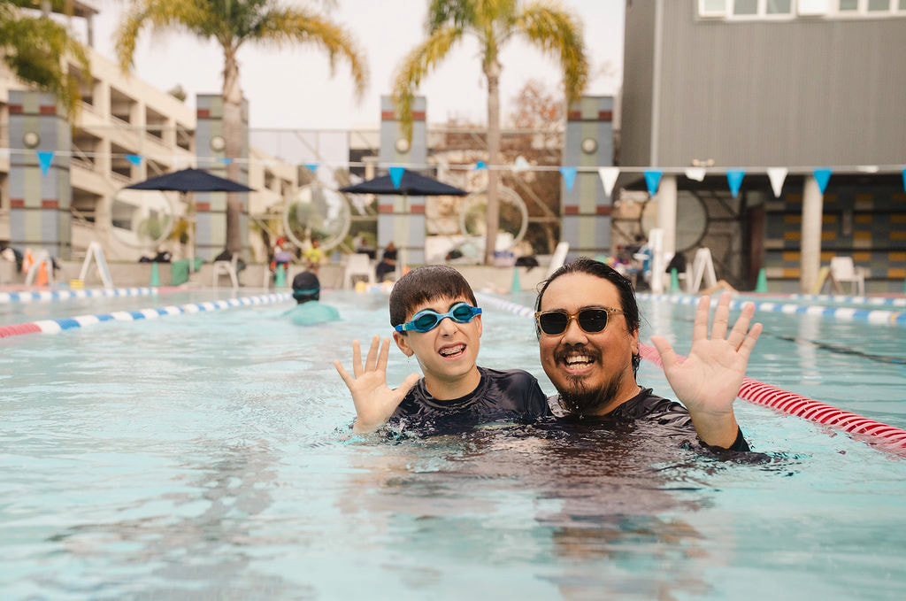 An instructor and a boy with blue goggles waving from an outdoor pool lane