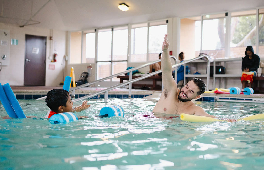 A bearded instructor demonstrating a backstroke arm movement to a child with foam noodles indoors