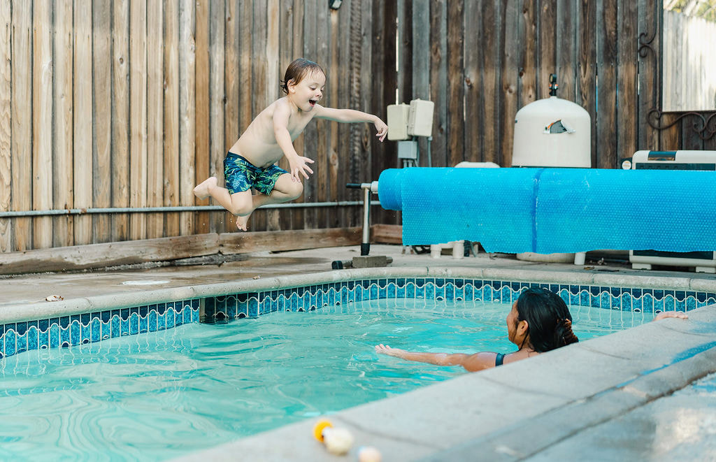 A young boy jumping into a backyard pool toward a female instructor waiting in the water
