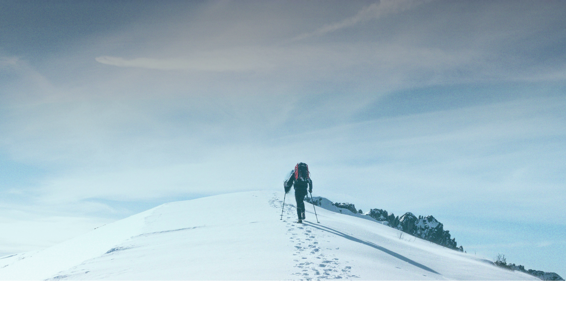 Lone hiker with trekking poles ascending a snowy mountain ridge under a pale blue sky