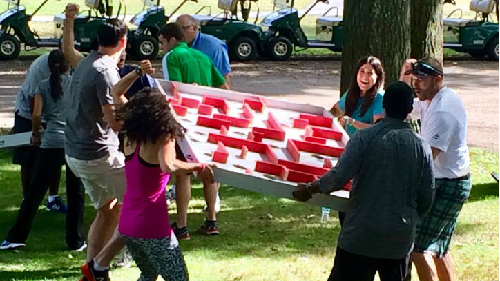 Team building participants carrying a large red-and-white puzzle board outdoors near golf carts