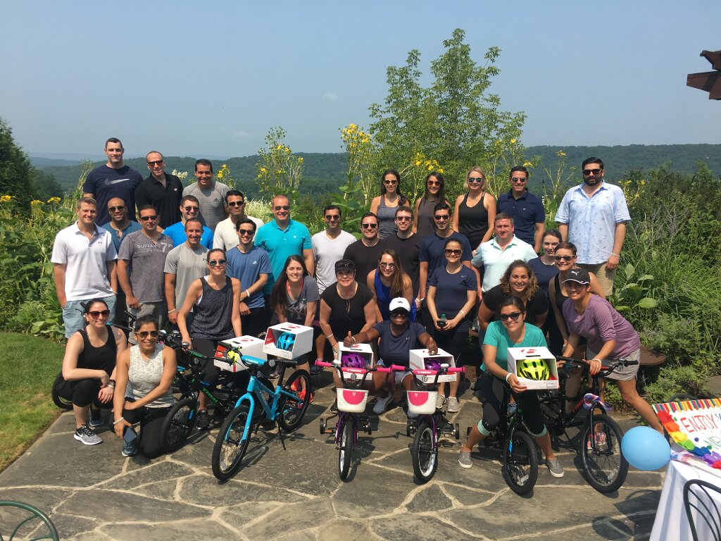 Large group of corporate employees posing outdoors with assembled charity bikes on a scenic hillside