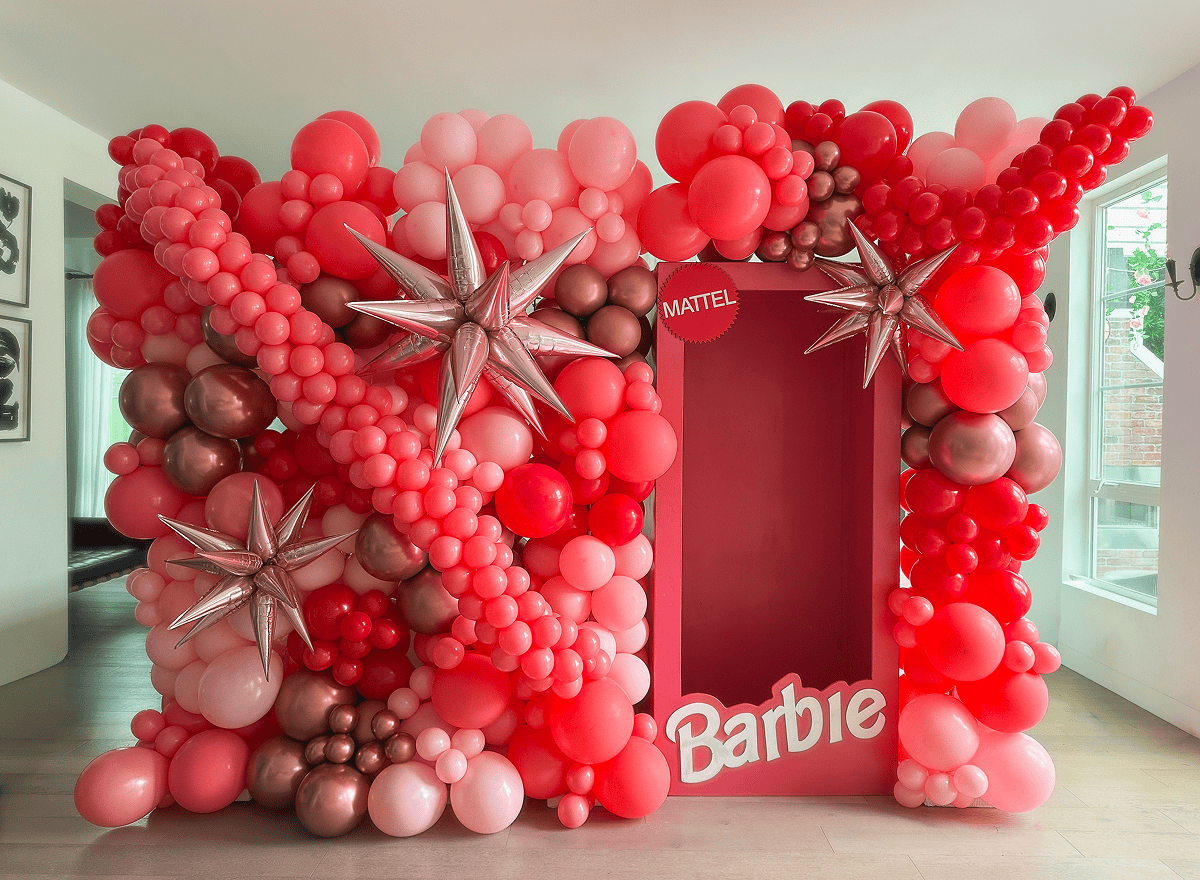 Red and pink balloon wall with a Barbie box prop and silver starburst foil balloons