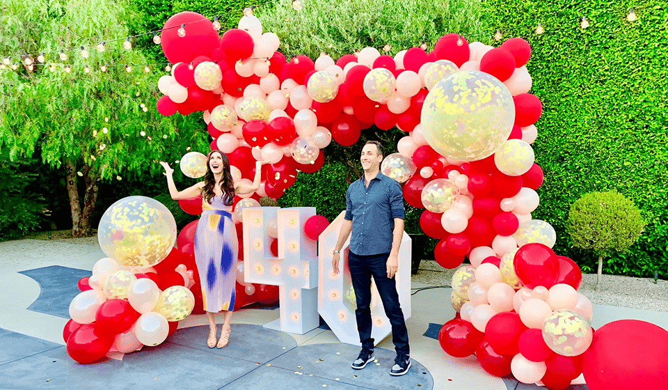 Couple in front of a large red and pink balloon garland arch with illuminated marquee letters