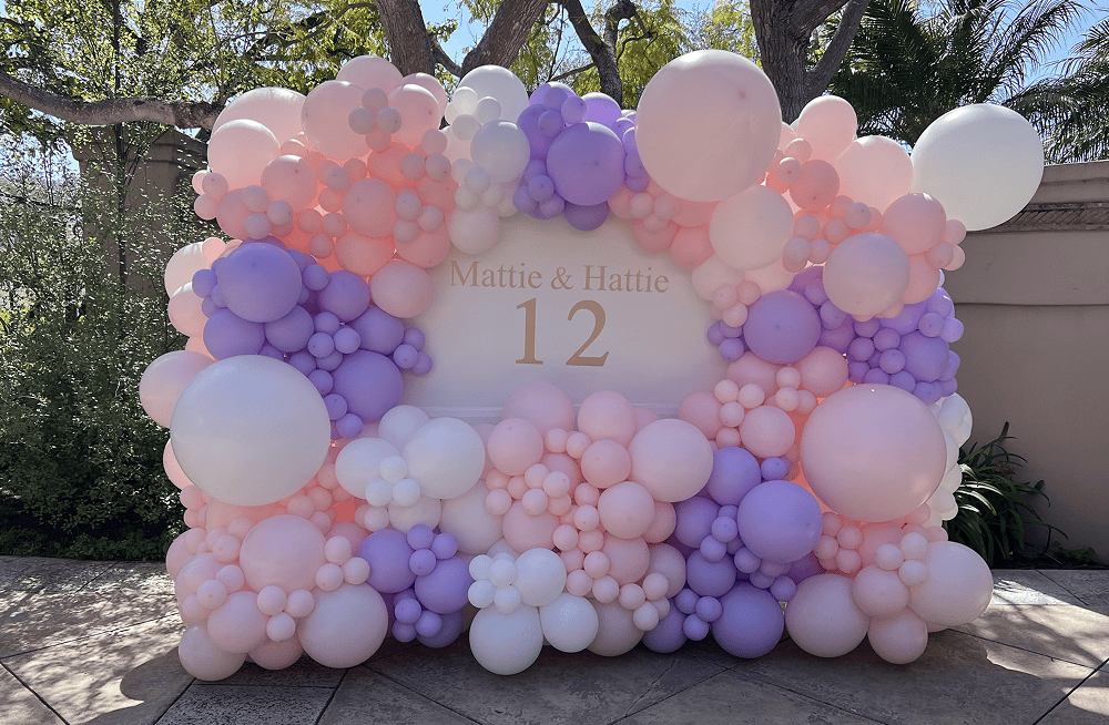 Pink, lavender and white balloon wall backdrop with a personalized "Mattie & Hattie 12" sign