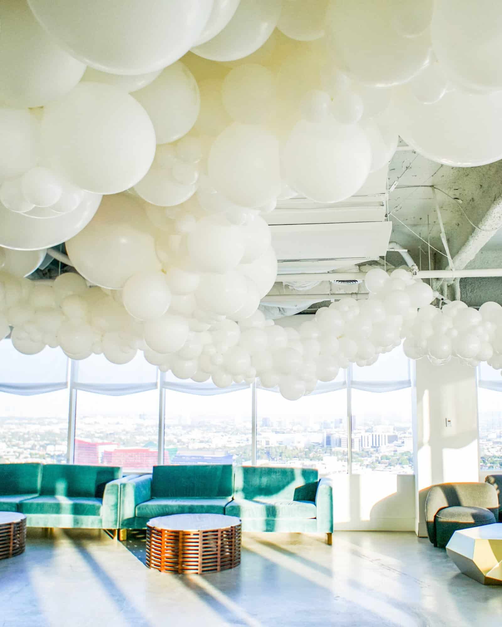 White balloon cloud ceiling installation over green velvet sofas at Soho House LA, with the city skyline through floor-to-ceiling windows