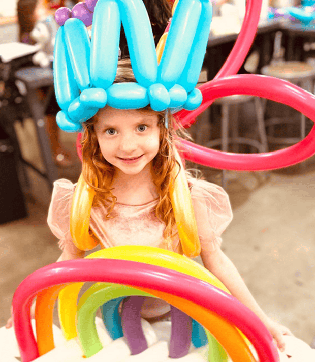 Girl wearing a blue twisted balloon crown holding a balloon rainbow sculpture