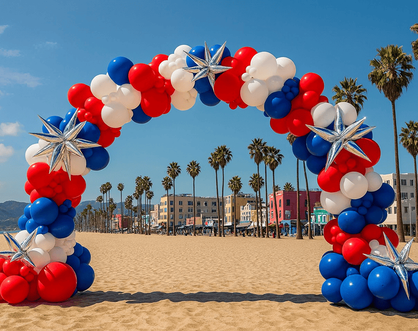 Red, white and blue patriotic balloon arch on a sandy beach with palm trees