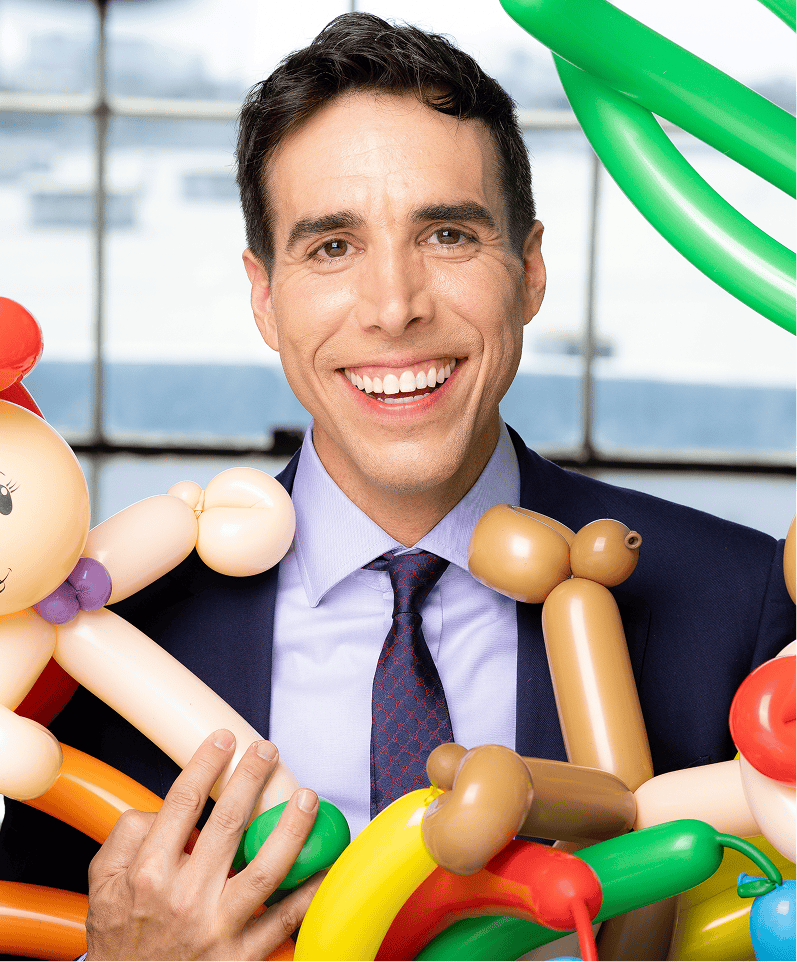 Sean, founder of The Balloon Guy, smiling in a suit while holding colorful balloon-animal sculptures with a city view behind him