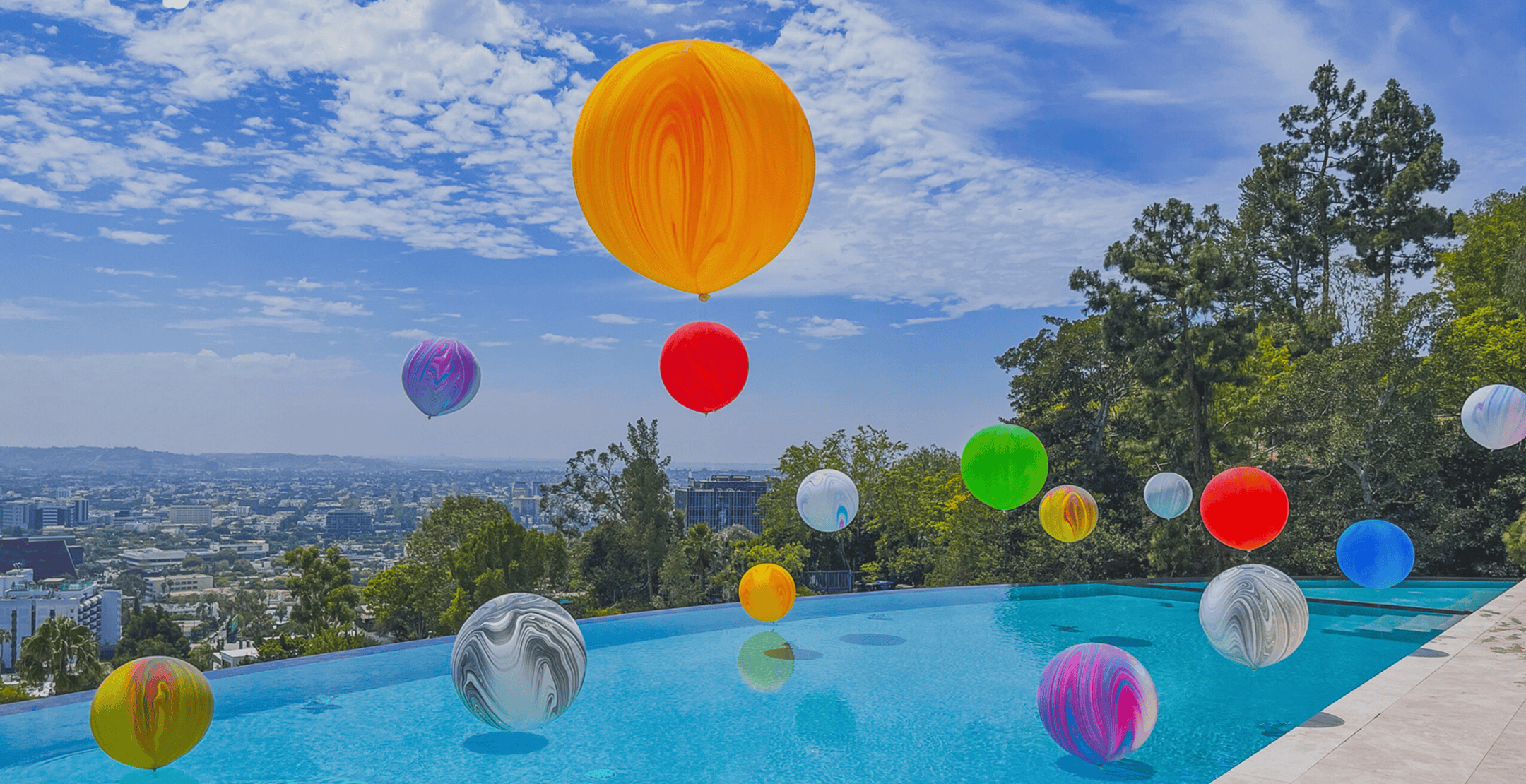 Large colorful marbled balloon spheres floating over an infinity pool with the Los Angeles cityscape behind
