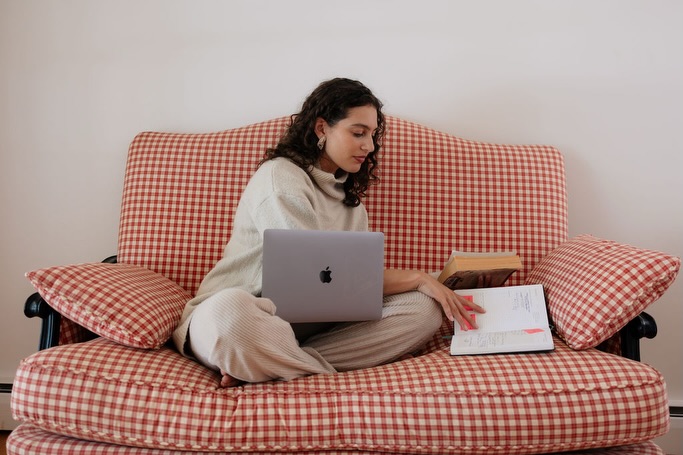 Chloe sitting cross-legged on a red gingham sofa working on a MacBook with a book in her lap