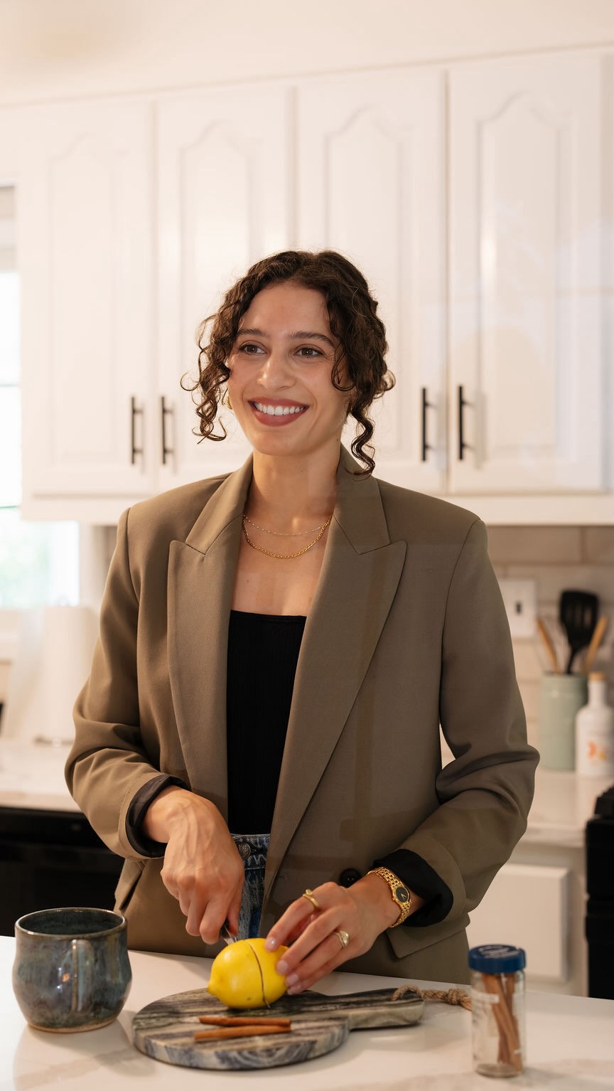 Chloe Sachs smiling in a kitchen wearing a khaki blazer, slicing a lemon beside a blue mug