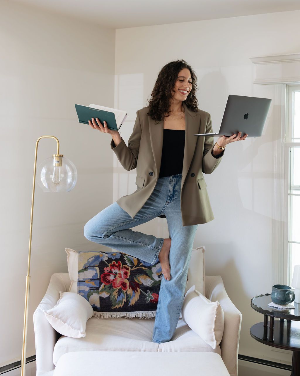 Chloe Sachs standing on a white armchair balancing a notebook and a laptop, wearing a khaki blazer and jeans