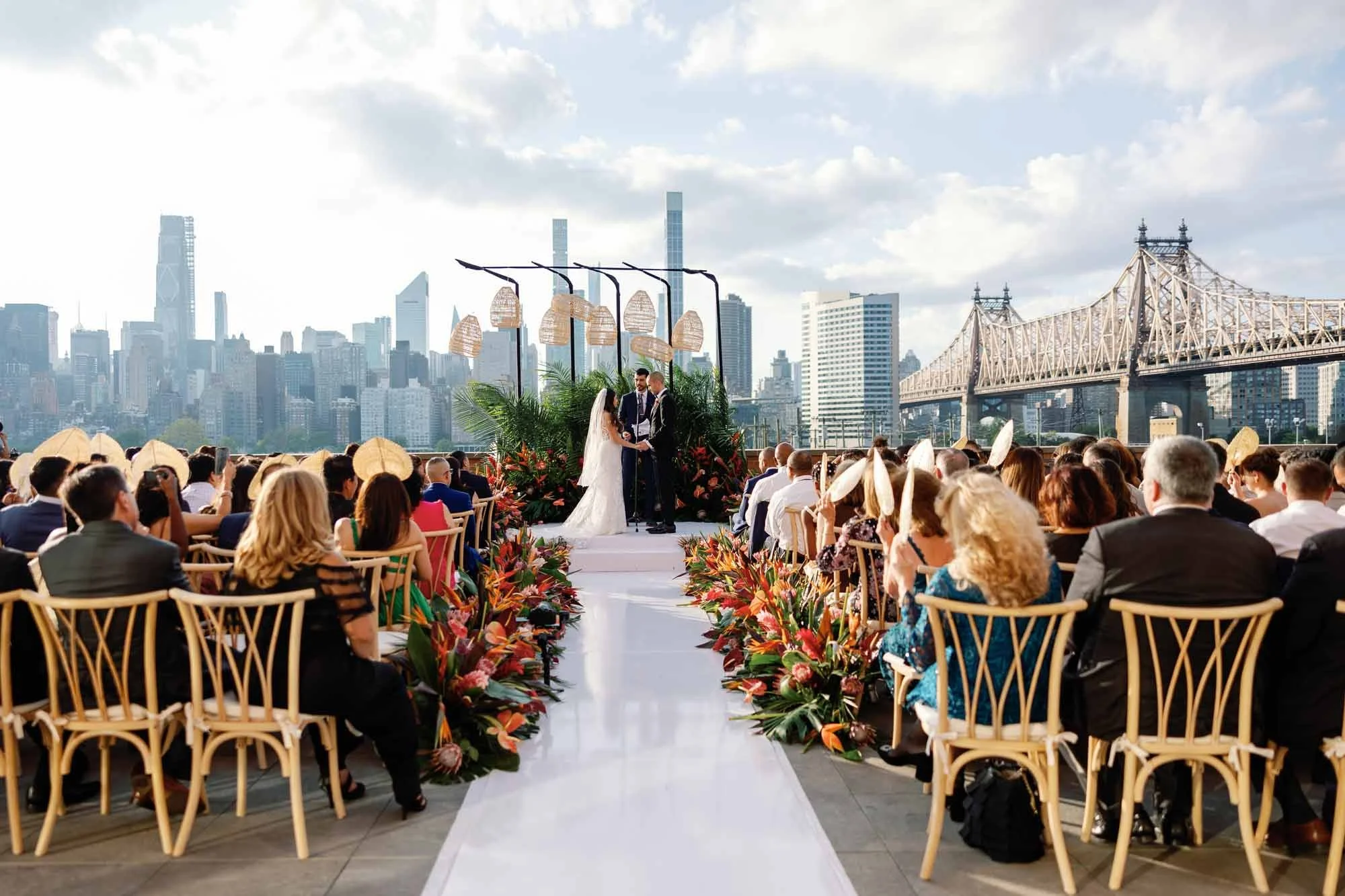 Outdoor rooftop wedding ceremony with tropical floral aisle, couple at altar, Queensboro Bridge and Manhattan skyline in background