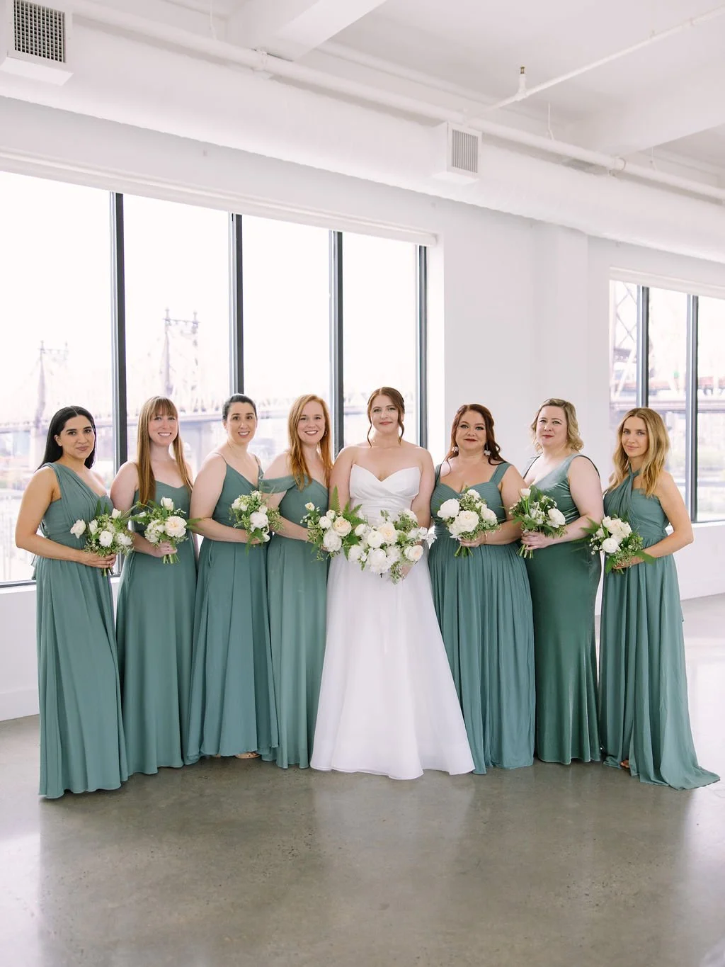 Bride flanked by seven bridesmaids in sage green dresses with Queensboro Bridge visible through windows