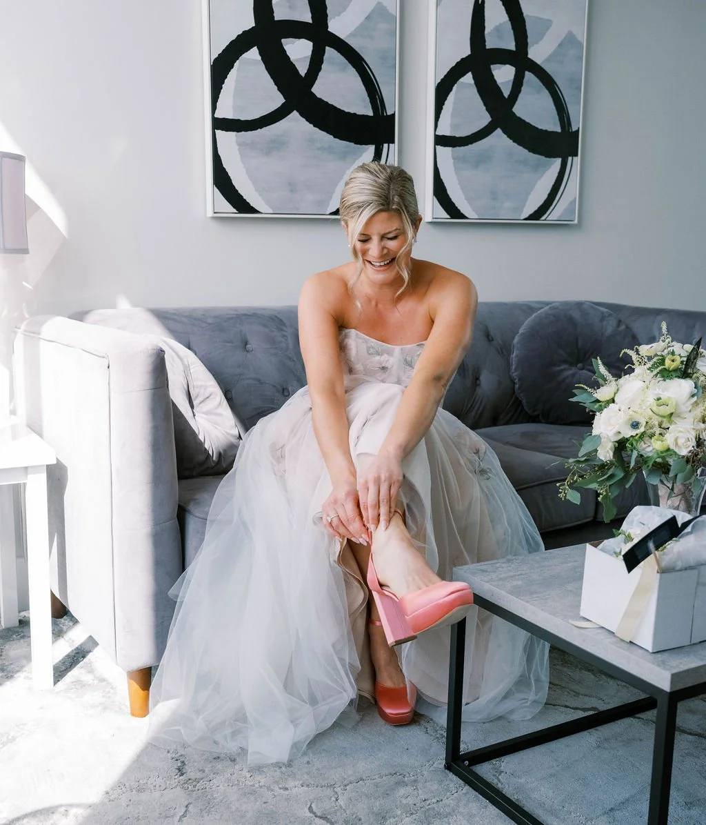 Bride in tulle gown putting on pink heels on a grey sofa in a modern bridal suite