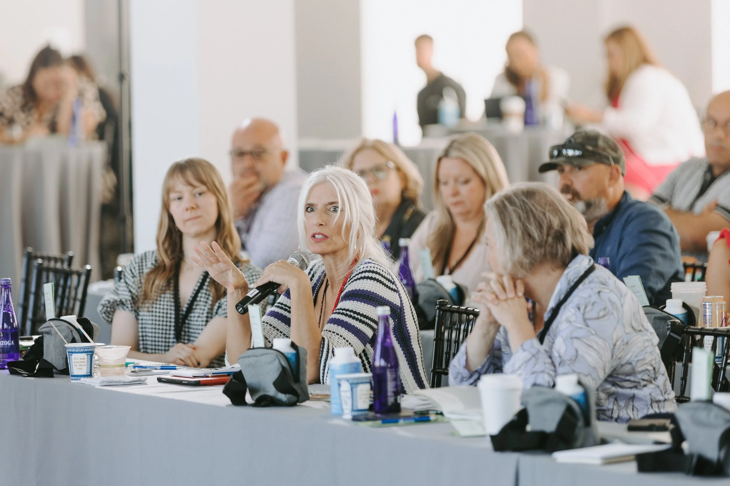Woman with platinum blonde hair speaking into a microphone at a conference table, attendees seated around her