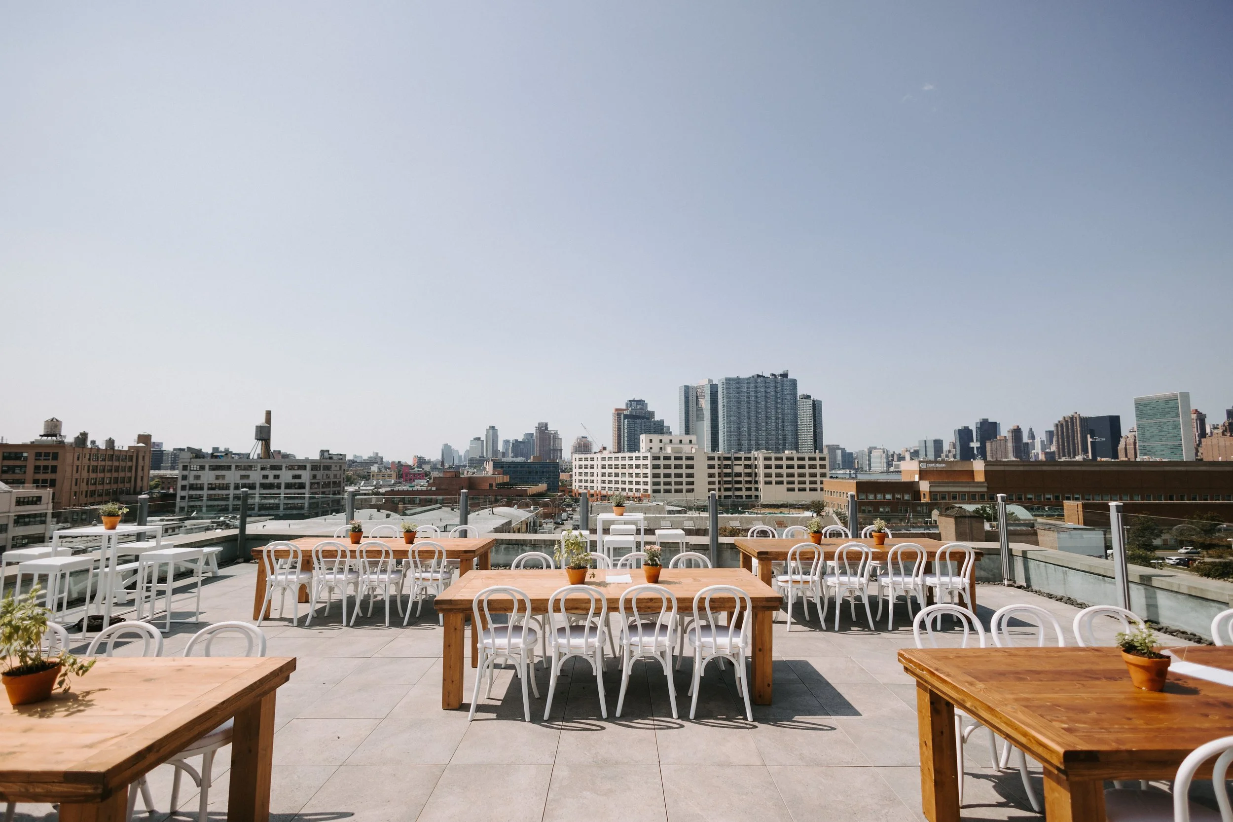 Rooftop terrace set with wooden farm tables and white chairs overlooking the NYC skyline