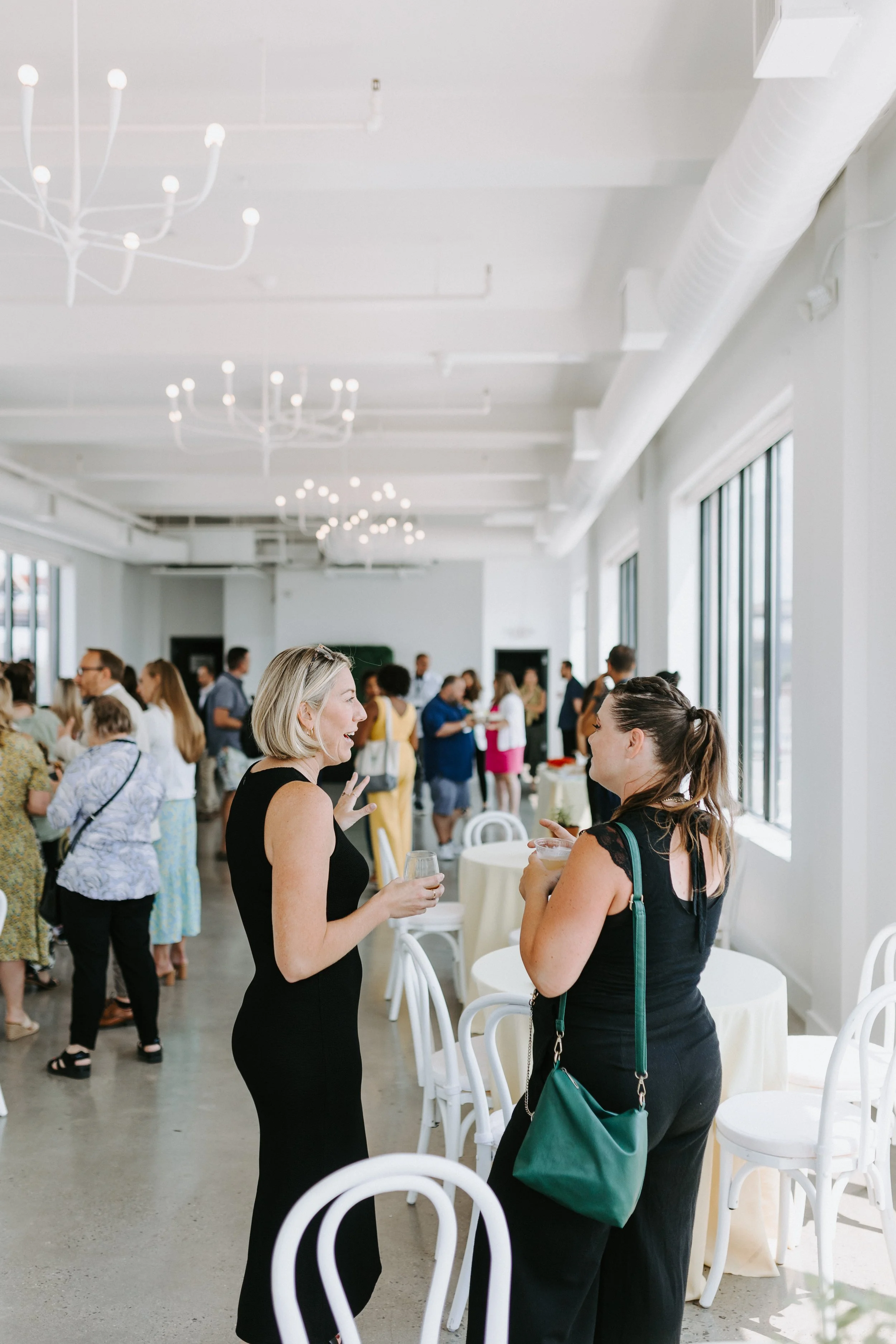 Two women in black dresses conversing at a networking event in a bright white event space with chandeliers
