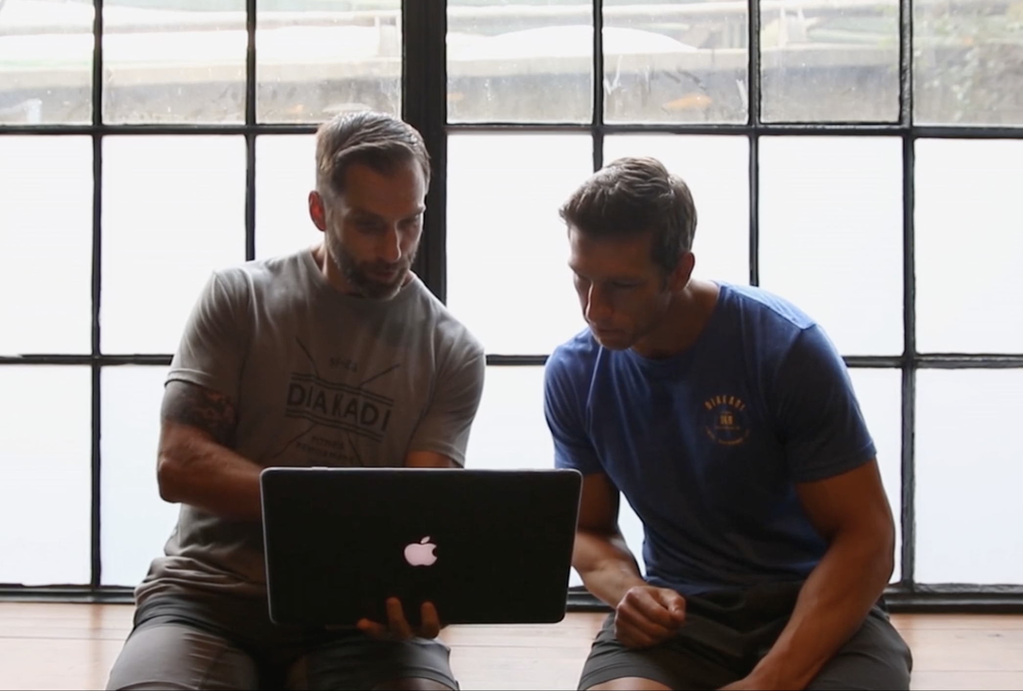 Two coaches in Diakadi-branded shirts reviewing a MacBook by warehouse windows