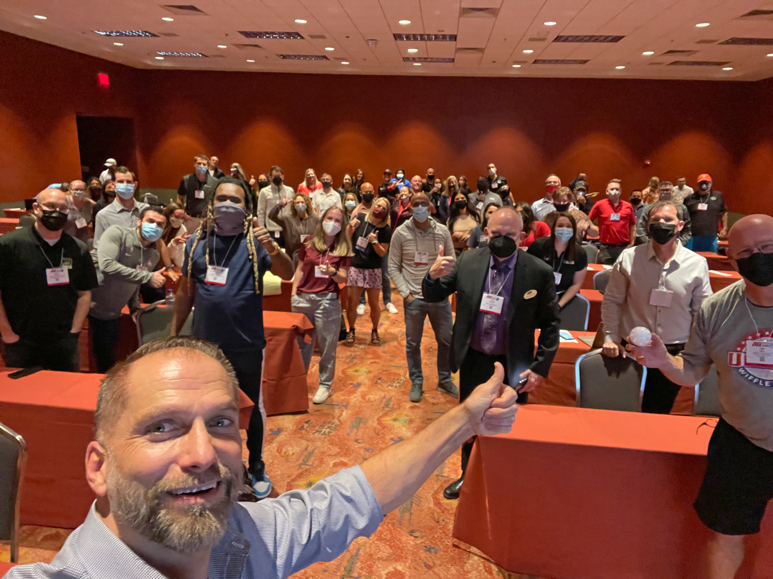 Billy Polson taking a thumbs-up selfie with a large conference audience at the Athletic Business Show